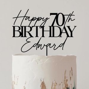 May include: A white frosted cake with gold and teal accents, topped with a black cake topper that reads "Happy 70th Birthday Edward." The cake sits on a wooden cake stand with a round top and pedestal base. The background is a neutral color.