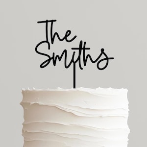 May include: A two-tiered white frosted cake on a wooden cake stand. A black cake topper reads "The Smiths" in a cursive font. The cake has a textured buttercream frosting.