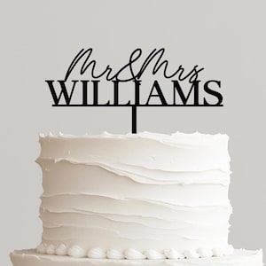 May include: A two-tiered white wedding cake with textured frosting, topped with a black cake topper that reads "Mr & Mrs WILLIAMS". The cake sits on a wooden cake stand.