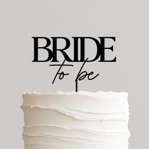 May include: A two-tiered white frosted cake on a wooden cake stand. A black cake topper reads "BRIDE to be". The cake has a textured frosting design and is ready for a wedding celebration.