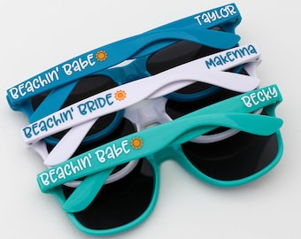 May include: A stack of sunglasses in shades of blue, white, and teal. Each pair has dark lenses and the text "BEACHIN' BABE", "BEACHIN' BRIDE", or a name printed on the arm. A small sun graphic is also present.