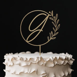 May include: A white cake with ruffled frosting and gold bead trim sits on a wooden cake stand. A gold-colored cake topper features the letter "G" inside a circle with a floral design. The cake is on a rustic wooden table.