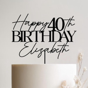 May include: A white cake with floral detailing and a cake topper that reads "Happy 40th Birthday Elizabeth." The cake is displayed on a wooden cake stand and decorated with dried flowers.