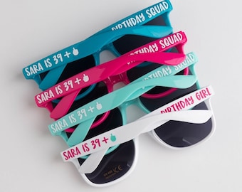 Peut inclure: Plusieurs paires de lunettes de soleil colorées, notamment turquoise, rose et blanche. Chaque paire a des verres foncés et des inscriptions sur les branches. Les inscriptions comprennent "SARA IS 39+", "BIRTHDAY SQUAD" et "BIRTHDAY GIRL".