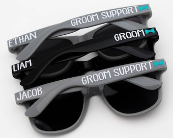 May include: Three pairs of gray sunglasses with black lenses. The top pair has the text "Groom Support" with a blue bowtie icon. The middle pair has the text "Groom" with a blue bowtie icon. The bottom pair has the text "Groom Support" with a blue bowtie icon. The sunglasses are stacked on top of each other. The top pair has the name "Ethan" on the left arm, the middle pair has the name "Liam" on the left arm, and the bottom pair has the name "Jacob" on the left arm.