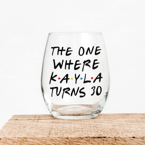 May include: A clear stemless wine glass with black text that reads "THE ONE WHERE KAYLA TURNS 30". The word "KAYLA" has the colors red, yellow, and blue. The glass is on a wooden surface.