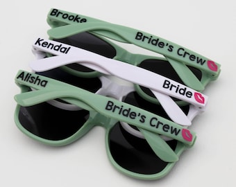 May include: Four pairs of green sunglasses with black text and pink lips. The sunglasses are stacked on top of each other. The text on the sunglasses reads "Bride's Crew", "Bride", "Kendal", and "Alisha".