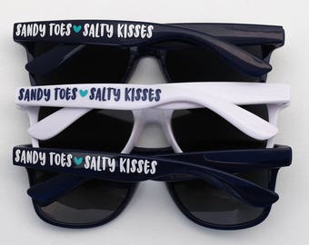 May include: Three pairs of sunglasses with black frames and dark lenses. The sunglasses have white text that reads "Sandy Toes Salty Kisses" on the top of the frames.