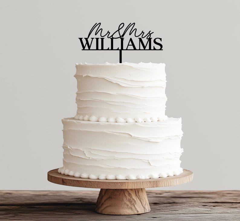 May include: A two-tiered white wedding cake with textured frosting, topped with a black cake topper that reads "Mr & Mrs WILLIAMS". The cake sits on a wooden cake stand.