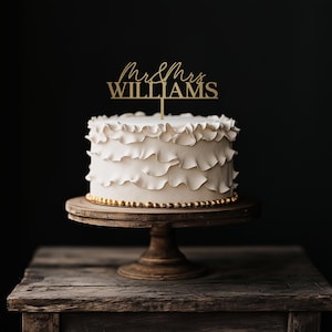 May include: A white wedding cake with ruffled frosting, displayed on a wooden cake stand. A gold-colored cake topper reads "Mr & Mrs WILLIAMS". The cake is decorated with gold-colored beads around the base, set against a dark background.