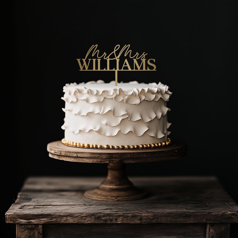 May include: A white wedding cake with ruffled frosting, displayed on a wooden cake stand. A gold-colored cake topper reads "Mr & Mrs WILLIAMS". The cake is decorated with gold-colored beads around the base, set against a dark background.