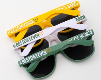 May include: Three pairs of sunglasses with black lenses and yellow, white, and green frames. The frames have the text "#BESTDAYEVER" and a heart symbol on them. The sunglasses also have the text "Bride's Babes", "Bride", and "Maid of Honor" on them.