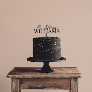 May include: A black cake with gold flecks on a black cake stand, topped with a black cake topper that reads "Mr & Mrs WILLIAMS". The cake sits on a wooden table against a neutral background.