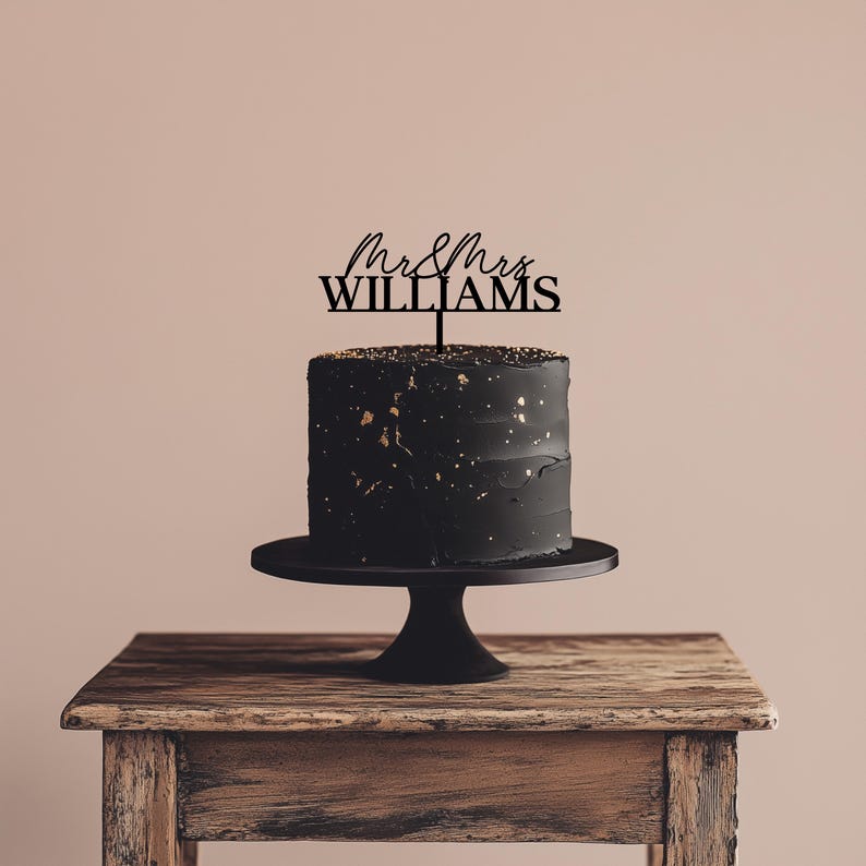May include: A black cake with gold flecks on a black cake stand, topped with a black cake topper that reads "Mr & Mrs WILLIAMS". The cake sits on a wooden table against a neutral background.