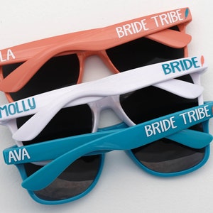 Peut inclure: Trois paires de lunettes de soleil avec des noms personnalis&eacute;s et la phrase "Bride Tribe" sur les branches. Les lunettes de soleil sont en corail, blanc et turquoise.