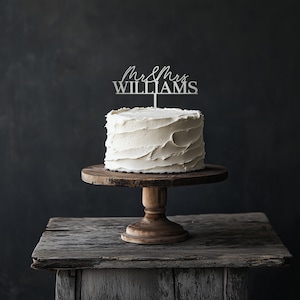 May include: A white frosted cake with a silver cake topper that reads "Mr & Mrs WILLIAMS" sits on a wooden cake stand. The cake stand is on a rustic wooden table against a dark background.