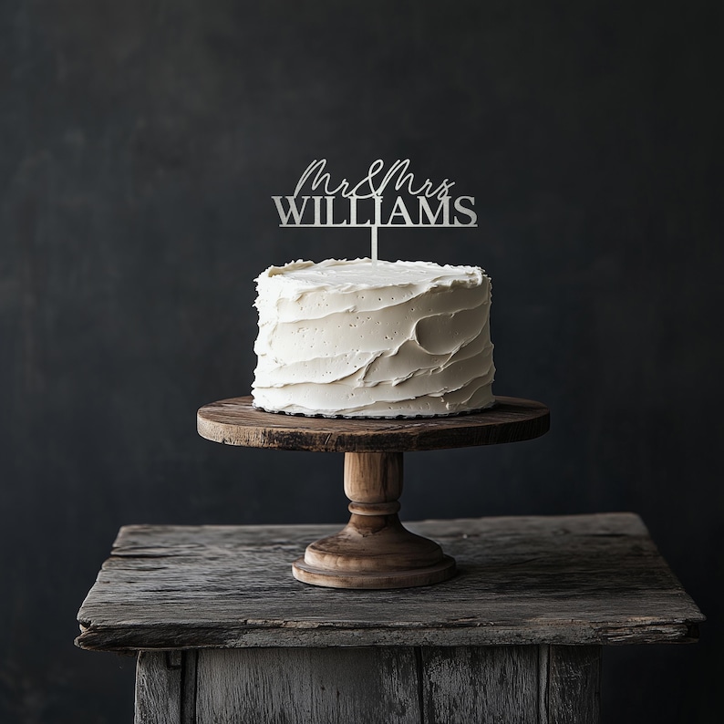 May include: A white frosted cake with a silver cake topper that reads "Mr & Mrs WILLIAMS" sits on a wooden cake stand. The cake stand is on a rustic wooden table against a dark background.