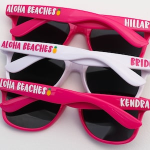 Peut inclure: Trois paires de lunettes de soleil roses et blanches avec des verres noirs. Les lunettes de soleil portent le texte "Aloha Beaches" avec un motif d'ananas sur le haut de la monture et le texte "Hillary", "Bride" et "Kendra" sur les branches.