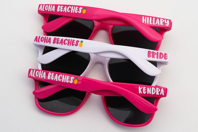 Peut inclure: Trois paires de lunettes de soleil roses et blanches avec des verres noirs. Les lunettes de soleil portent le texte "Aloha Beaches" avec un motif d'ananas sur le haut de la monture et le texte "Hillary", "Bride" et "Kendra" sur les branches.