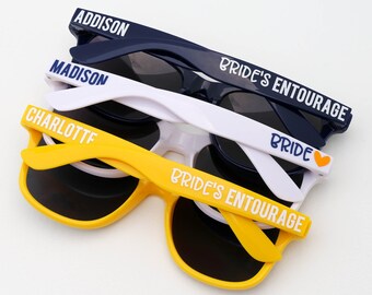 May include: Three pairs of sunglasses with black lenses. The top pair is navy blue with the text "ADDISON" on the frame. The middle pair is white with the text "MADISON" on the frame. The bottom pair is yellow with the text "CHARLOTTE" on the frame. All three pairs have the text "PRIDE'S  ENTOURAGE" on the frame. The middle pair also has the text "BRIDE" with a heart on the frame.