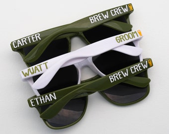 May include: Four pairs of sunglasses with different names on them. The sunglasses are green and white. The names on the sunglasses are "Brew Crew", "Groom", "Wyatt", "Carter", "Ethan", and "Brew Crew".