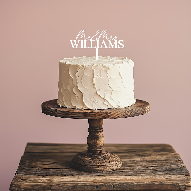 May include: A white frosted cake with a decorative topper that reads "Mr & Mrs Williams" sits on a wooden cake stand. The cake stand is placed on a rustic wooden table against a pink background. The cake has a textured frosting.