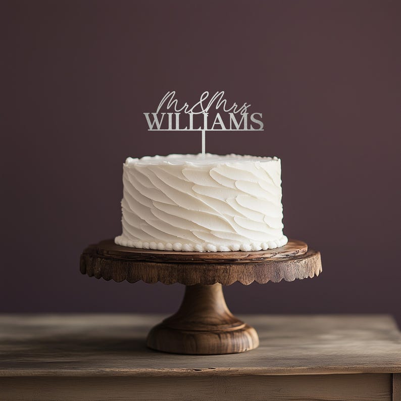 May include: A white wedding cake with textured frosting sits on a wooden cake stand. A silver cake topper reads "Mr & Mrs Williams". The cake stand is made of dark wood and has a scalloped edge.