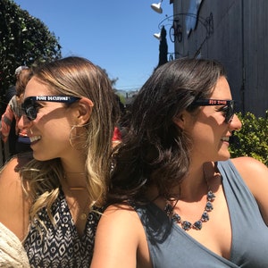 Peut inclure: Deux femmes portant des lunettes de soleil noires avec du texte blanc. Les lunettes de soleil &agrave; gauche disent "FOUR DECISIONS" et les lunettes de soleil &agrave; droite disent "SLO 2018".