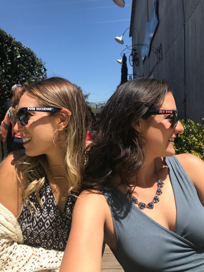 Peut inclure: Deux femmes portant des lunettes de soleil noires avec du texte blanc. Les lunettes de soleil &agrave; gauche disent "FOUR DECISIONS" et les lunettes de soleil &agrave; droite disent "SLO 2018".