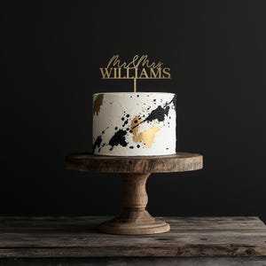 May include: A white cake with black and gold paint splatter design, topped with a gold "Mr & Mrs WILLIAMS" cake topper. The cake sits on a wooden cake stand, perfect for a wedding or anniversary celebration.