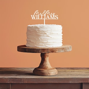 May include: A white frosted cake with a wooden cake stand. The cake is topped with a white cake topper that reads "Mr & Mrs WILLIAMS" in a script font. The cake stand is made of wood and has a pedestal base.