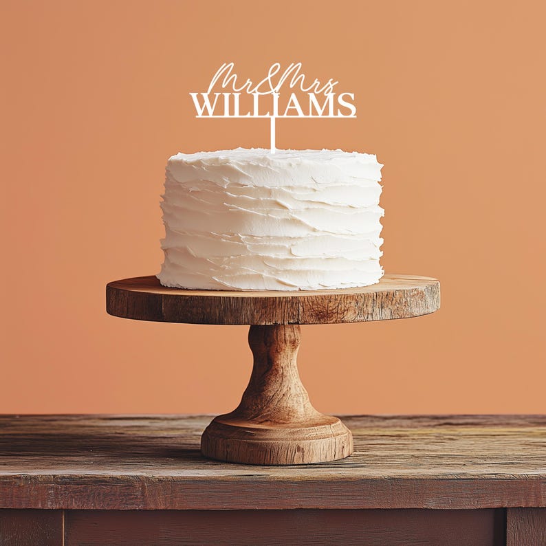 May include: A white frosted cake with a wooden cake stand. The cake is topped with a white cake topper that reads "Mr & Mrs WILLIAMS" in a script font. The cake stand is made of wood and has a pedestal base.