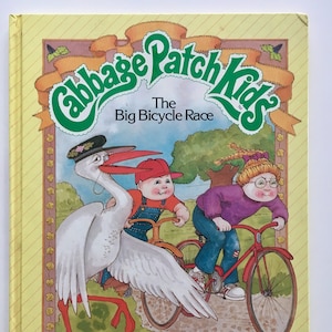 Pode incluir: Uma capa de livro amarela com um logotipo verde Cabbage Patch Kids e o título "The Big Bicycle Race". A capa apresenta uma ilustração de três Cabbage Patch Kids andando de bicicleta com uma cegonha branca.