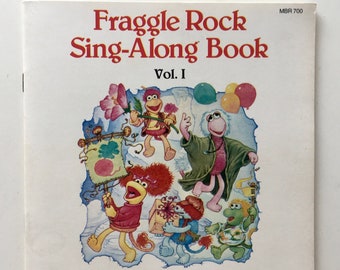 May include: A colorful book cover featuring the Fraggle Rock characters. The title is "Fraggle Rock Sing-Along Book Vol. I". The cover features illustrations of the Fraggles, including Red, Gobo, and Wembley, along with the text "SEE the pictures HEAR the story READ the book".