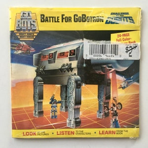 May include: A GoBots toy packaging featuring a large, grey robot with a red laser beam. The robot is standing on a gridded surface with other smaller robots. The packaging has the text "Challenge of the GoBots" and "Battle for GoBotron" in yellow letters. The packaging also includes a 24-page full-colour book.