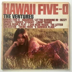 Könnte beinhalten: Vintage Vinyl-Schallplattenhülle für "Hawaii Five-O" von The Ventures. Das Cover zeigt eine Frau in einem geblümten Kleid, die im Gras liegt. Der Albumtitel und der Künstlername sind in roter und schwarzer Schrift. Songtitel sind unten aufgeführt.