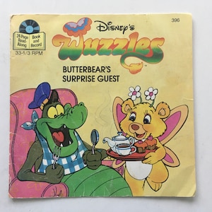 Könnte beinhalten: Ein Vintage-Buch und eine Schallplatte von Disney's Wizzles mit dem Titel "Butterbears Überraschungsgast". Das Cover zeigt eine grüne Krokodilfigur mit einem blauen Hut und eine gelbe Bärenfigur mit rosa Schmetterlingsflügeln, die ein Tablett mit Teetassen und Cupcakes hält.