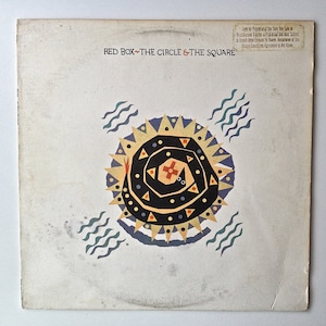 May include: A white vinyl record album cover with the title "Red Box - The Circle & The Square" printed in black and red. The cover features a colorful illustration of a sun with a black and yellow spiral design.