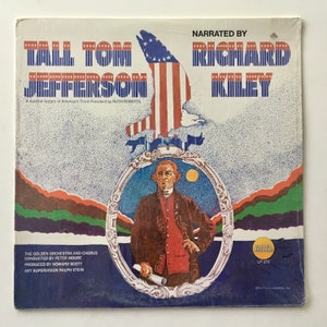 以下が含まれることがあります： アメリカの3代目大統領トーマス・ジェファーソンを描いた、ビンテージのビニールレコードアルバムのカバー。カバーには、ハクトウワシとアメリカの国旗が描かれています。アルバムのタイトルは「トール・トム・ジェファーソン」で、リチャード・カイリーがナレーションを担当しています。このアルバムは、トーマス・ジェファーソンの生涯を描いた音楽史です。
