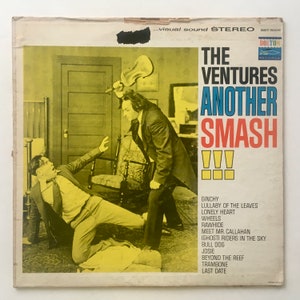 Op de afbeelding: Een vintage vinyl platenhoes met een foto van twee mannen in een kamer. De hoes is verkleurd door de tijd en heeft de titel "The Ventures Another Smash!!!" in rood, blauw en wit. Het platenlabel is Dolton Records.