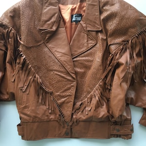 May include: A brown leather jacket with fringe detailing. The jacket has a zipper closure and a snap button closure. The label on the jacket reads "LEMONT".