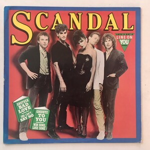 May include: A group of five people, a band called "SCANDAL", posing for a photo. The band members are wearing black, white, and grey clothing. The background is red. The album cover features the title "LOVE'S GOT A LINE ON YOU" and the names of some of the band's songs: "Another Bad Love", "She Can't Say No", "Goodbye To You", and "Win Some, Lose Some".