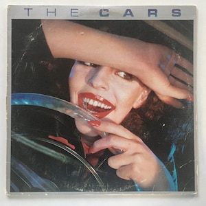 May include: The Cars album cover featuring a woman with red lipstick and a mischievous smile, holding a steering wheel. The album title "THE CARS" is printed in silver letters at the top of the cover.
