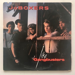 May include: A record album cover featuring a band called "Jo Boxers" with the title "Like Gangbusters" in red text. The cover features five young men in casual clothing standing in front of a wooden structure.