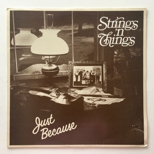Könnte beinhalten: Ein sepiafarbenes Albumcover mit einer Vintage-Öllampe, einem gerahmten Foto und einer Geige. Die Texte "Strings 'n Things" und "Just Because" sind in eleganter Schrift dargestellt. Das Thema deutet auf eine musikalische oder Vintage-Ästhetik hin.