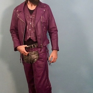 May include: A purple leather jacket with a zipper closure and a matching purple leather chaps. The jacket has a stand-up collar and a zippered pocket on the left chest.