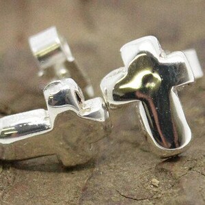 Puede incluir: Un par de pendientes de bot&oacute;n con forma de cruz en tono plateado. Cada pendiente presenta una forma de cruz lisa y redondeada con un acabado met&aacute;lico reflectante y un cierre de poste est&aacute;ndar, dise&ntilde;ado para orejas perforadas.