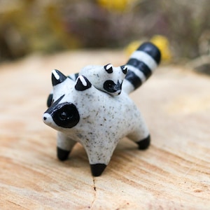 May include: A small, speckled ceramic raccoon figurine with a baby raccoon riding on its back. The raccoon has black markings around its eyes, ears, and feet, and a striped tail. The figurine is set on a wooden surface.