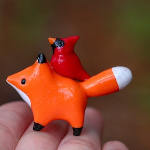 K&ouml;nnte beinhalten: Eine kleine orangefarbene Fuchsfigur mit einer wei&szlig;en Spitze am Schwanz, die eine rote Kardinalfigur auf dem R&uuml;cken tr&auml;gt.