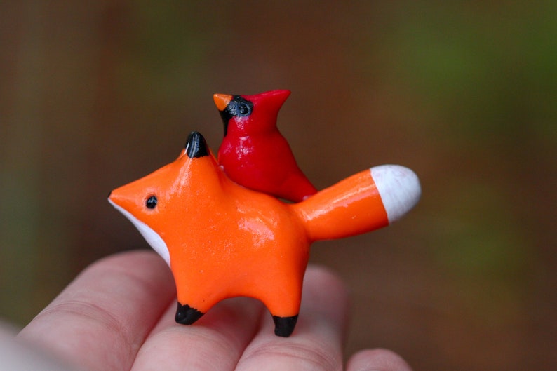 K&ouml;nnte beinhalten: Eine kleine orangefarbene Fuchsfigur mit einer wei&szlig;en Spitze am Schwanz, die eine rote Kardinalfigur auf dem R&uuml;cken tr&auml;gt.